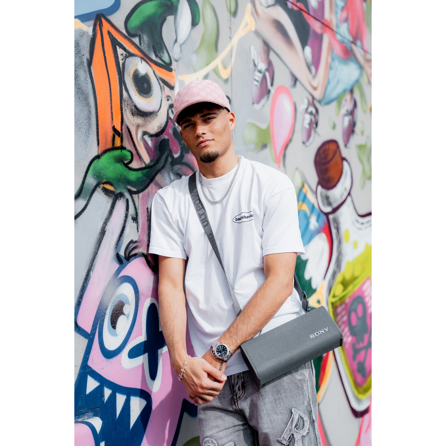Man leunt op graffiti-muur, draagt roze hoed en wit shirt. Een grijze Sony-luidsprekertas hangt.