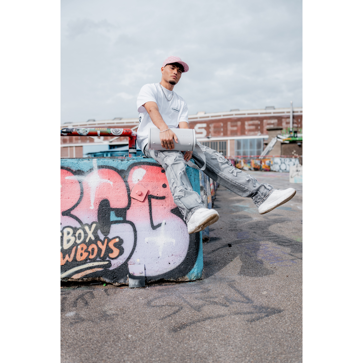 Un homme est assis sur un mur de graffitis, portant une casquette rose et des vêtements blancs, tenant un sac.