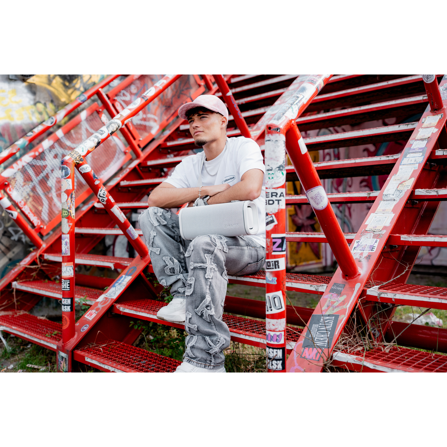 Homme avec enceinte Sony blanche, assis sur escalier rouge graffité. Casquette rose.