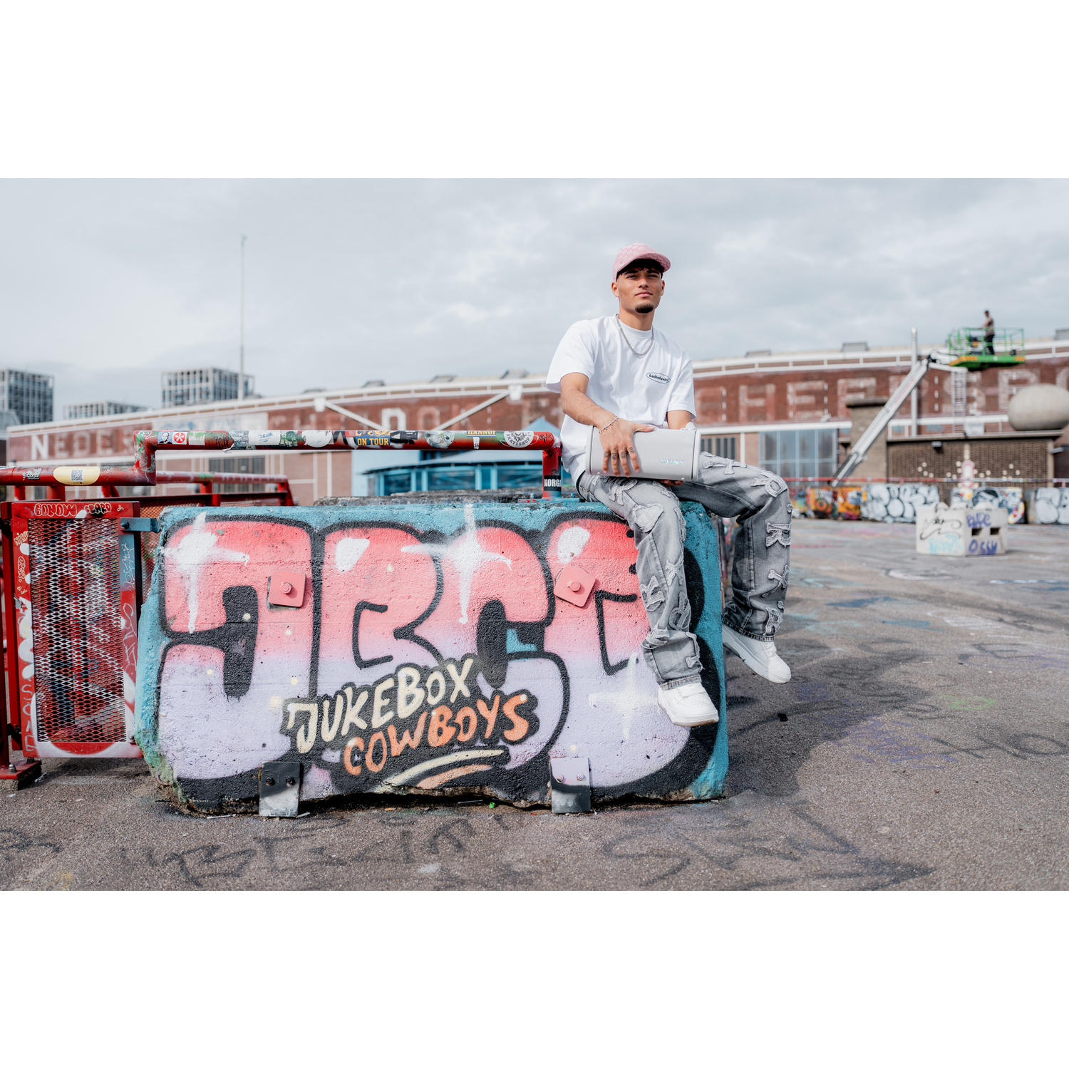 Un homme assis sur un bloc de béton couvert de graffitis, tenant un ordinateur portable, en extérieur.