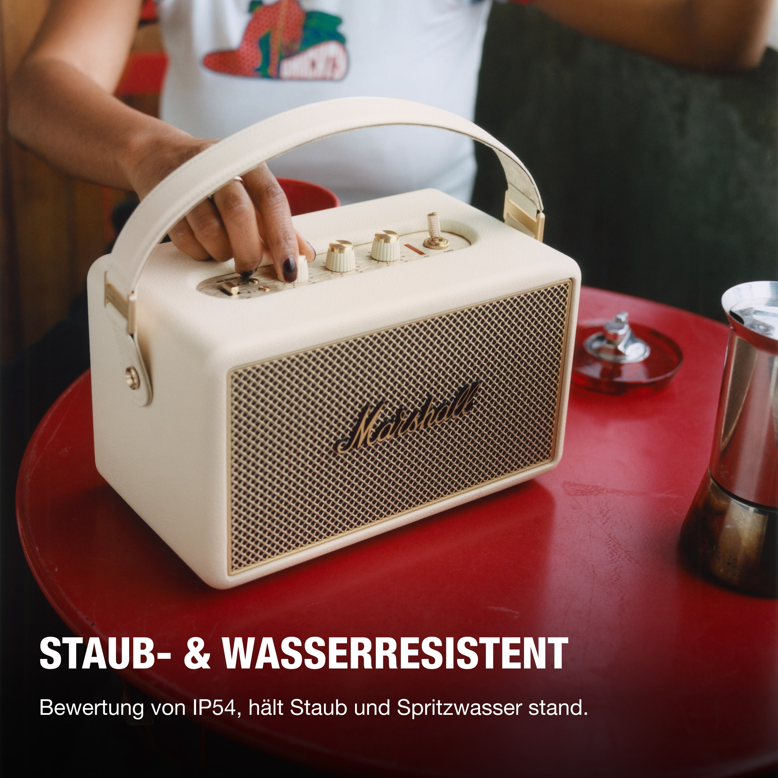 Cremefarbener Marshall-Lautsprecher auf rotem Tisch. Hand einer Frau stellt Knopf ein. Text: STAUB- & WASSERRESISTENT.