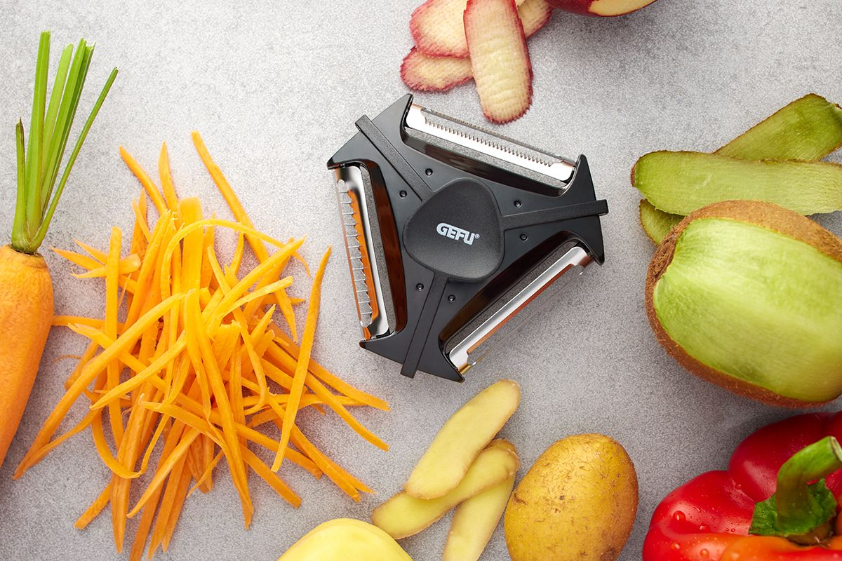 Gemüseschäler mit Karotten, Kiwi, Kartoffeln und Paprika. Der Schäler ist schwarz und hat mehrere Klingen.