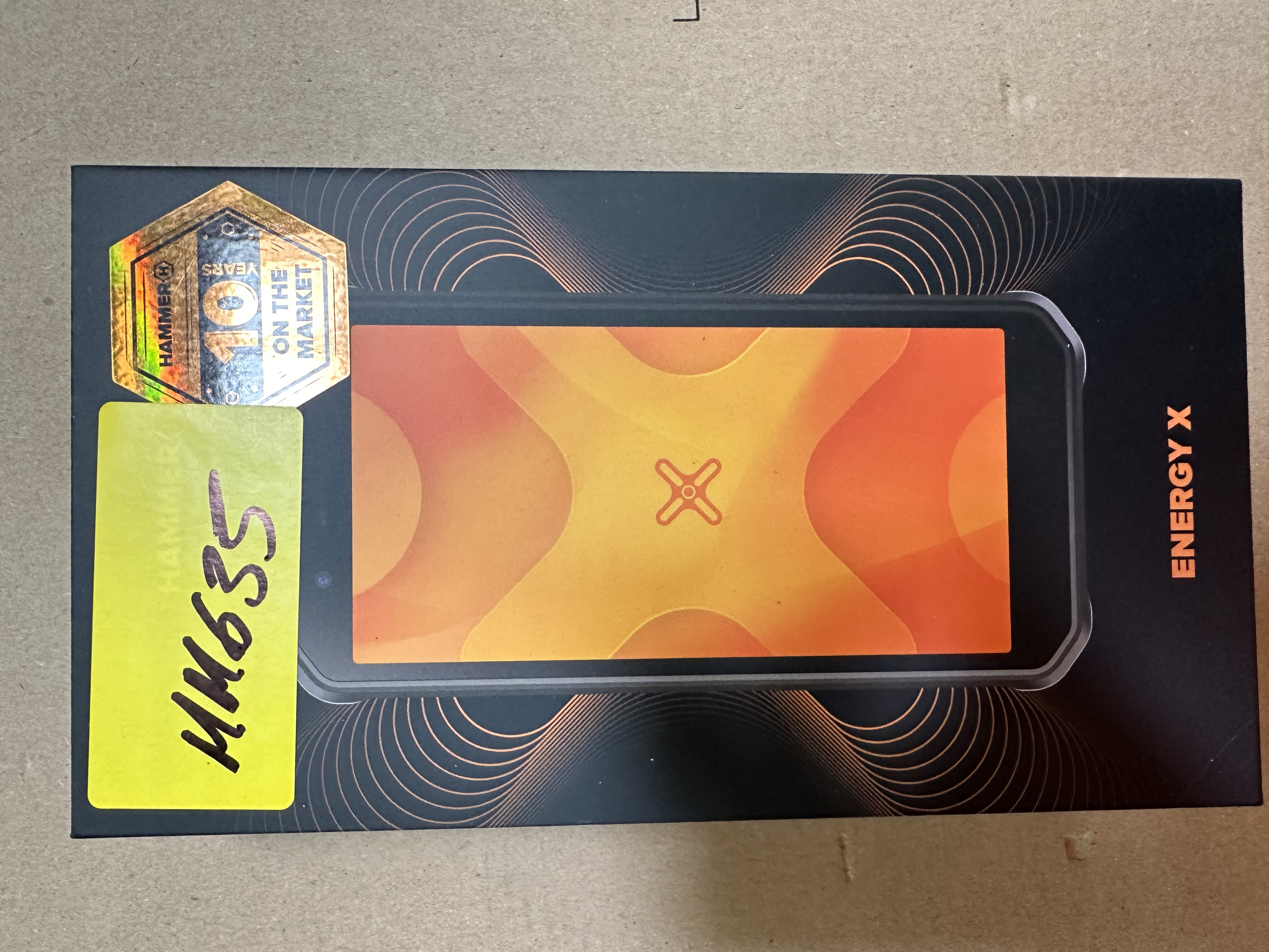 Czarny telefon w pudełku. Pokazuje ekran telefonu i logo. Żółta naklejka.