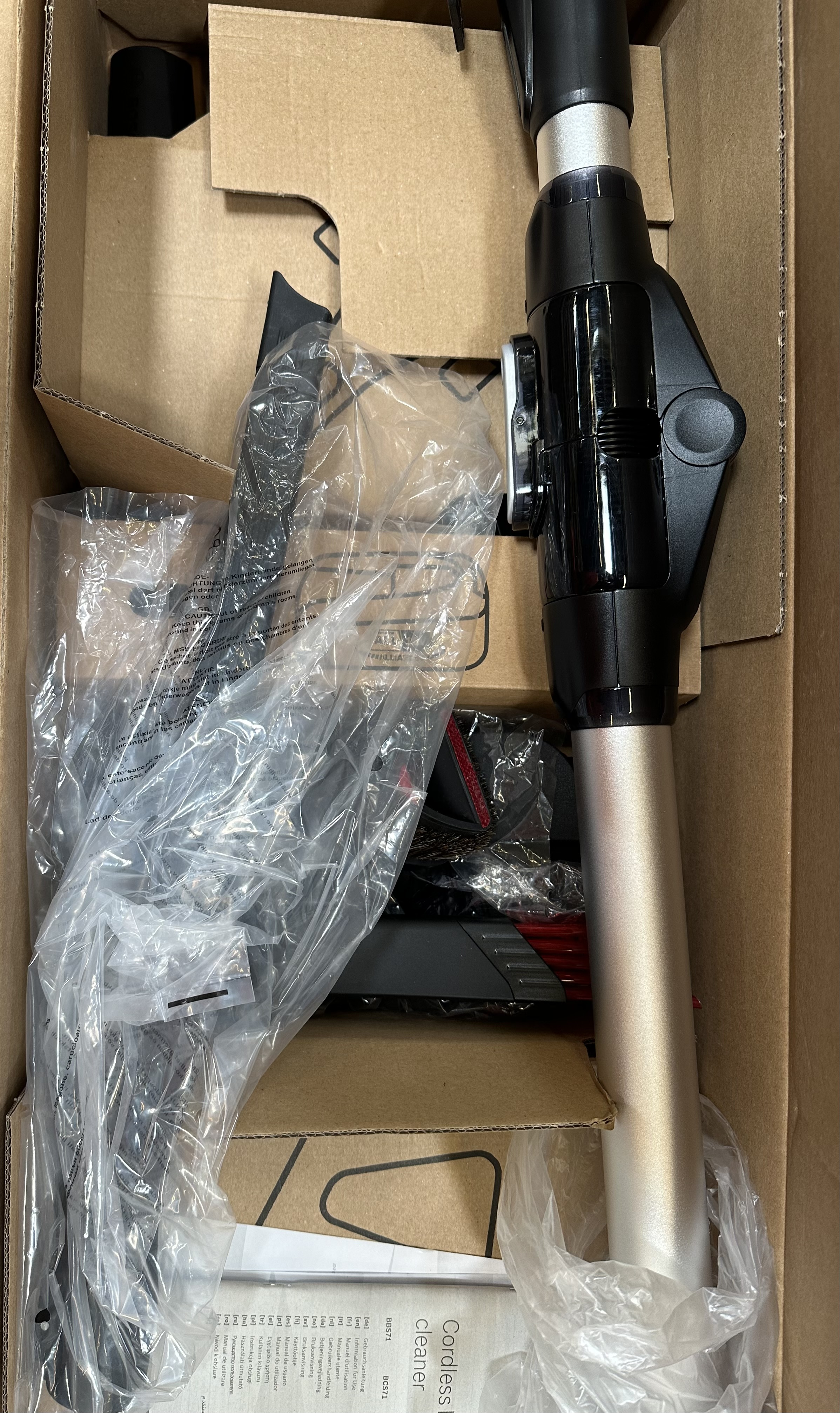 Części odkurzacza w kartonie. Głównie czarne i srebrne elementy.