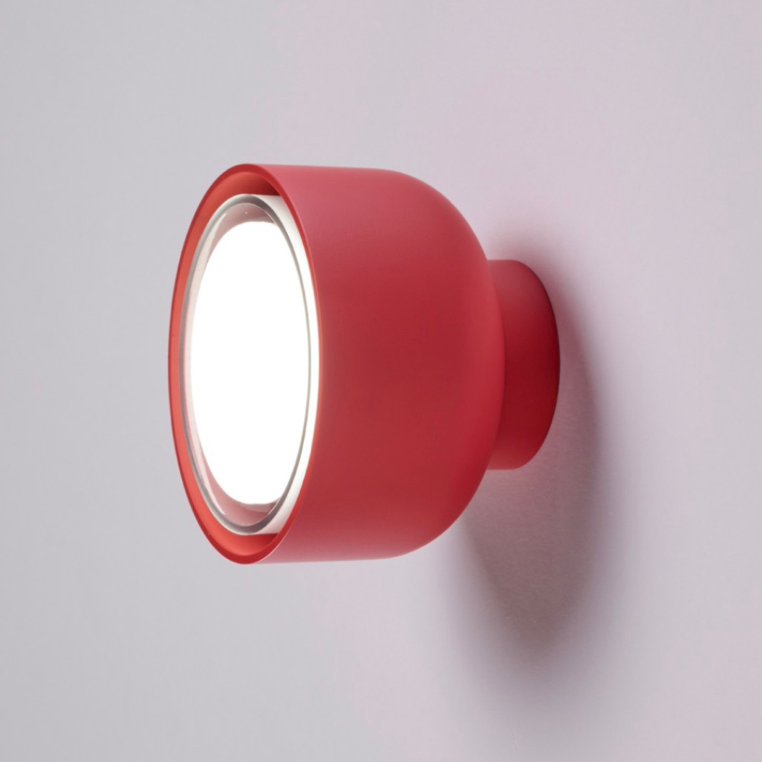 Luce da parete rossa e arrotondata con una sorgente luminosa circolare brillante. Montata su un muro bianco.