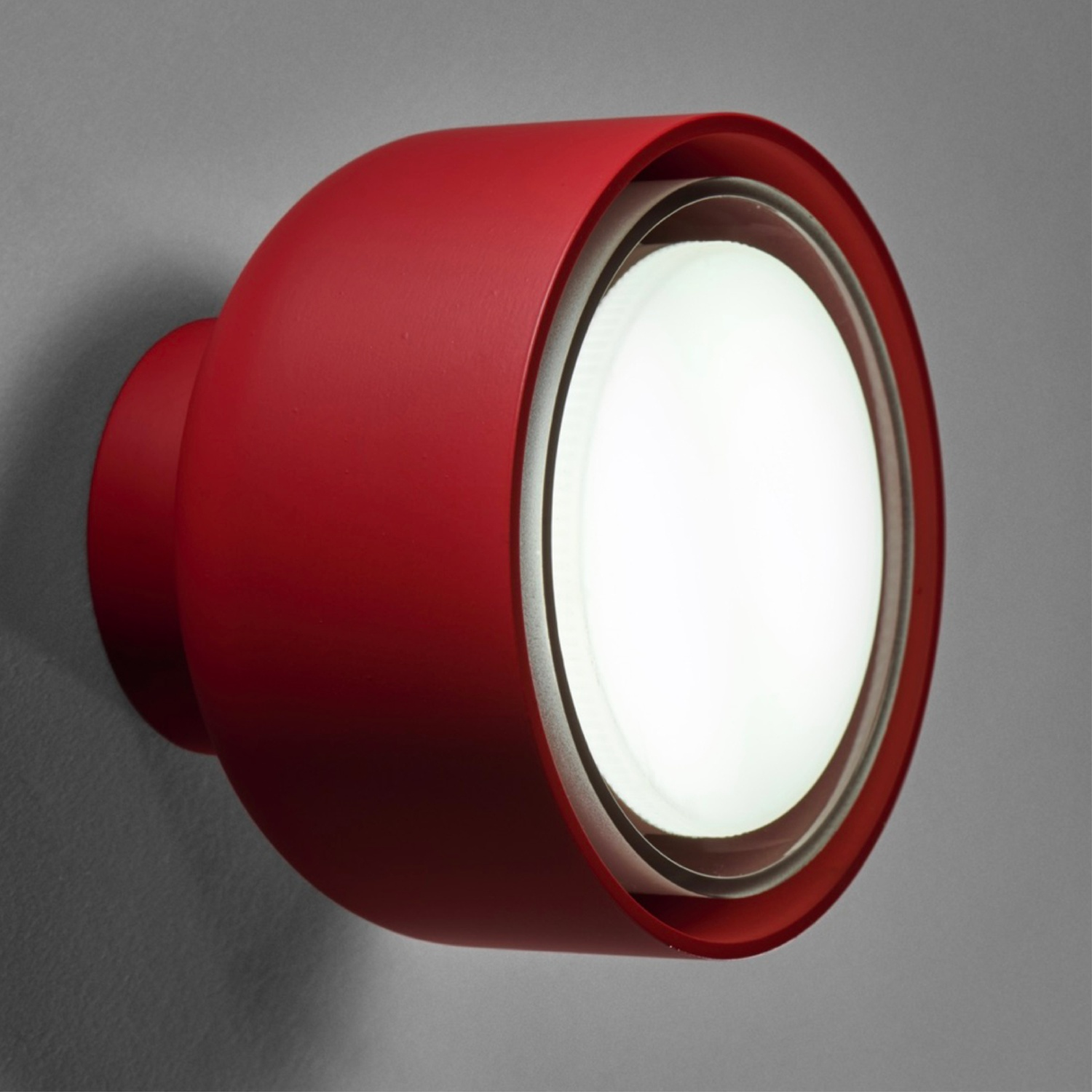 Lampada da parete rossa, rotonda e illuminata su muro grigio.