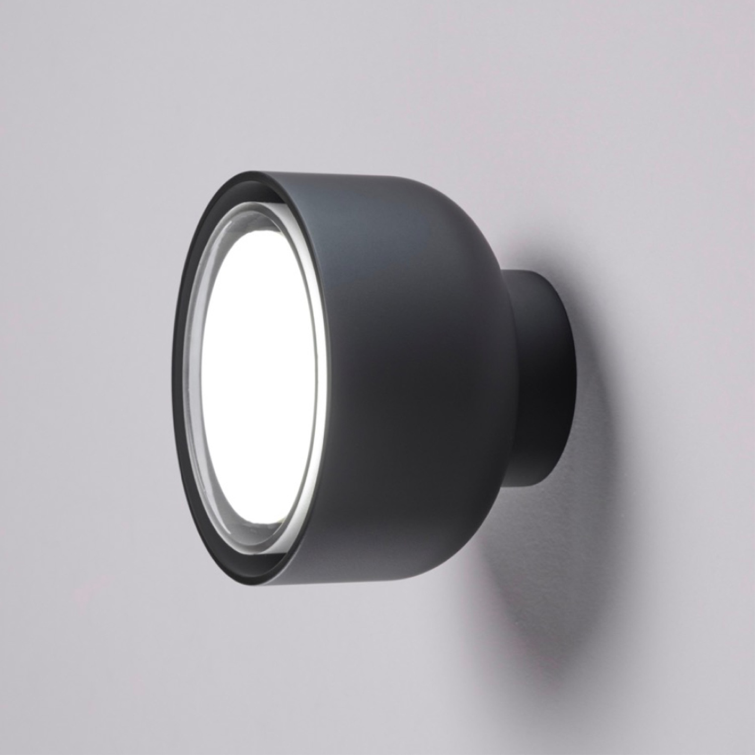 Un'applique nera con luce centrale brillante. Lo sfondo è un muro grigio.