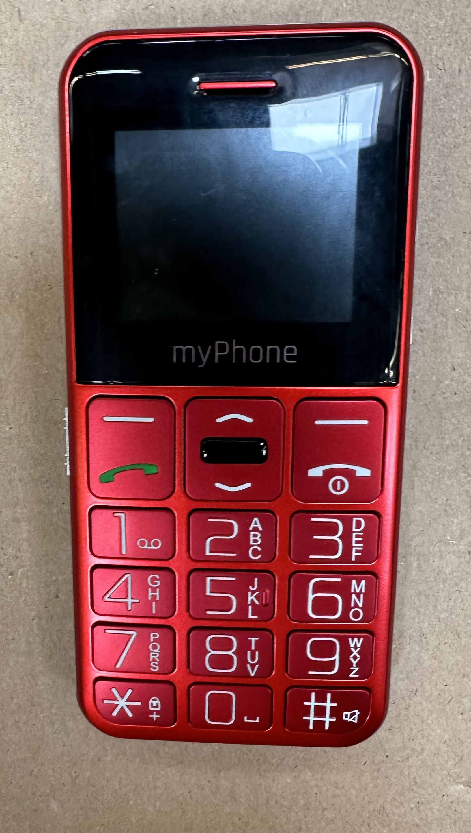 Czerwony telefon komórkowy myPhone z dużymi przyciskami. Czarny ekran, przyciski połączeń i wybierania. Tło to jasnobrązowa powierzchnia.
