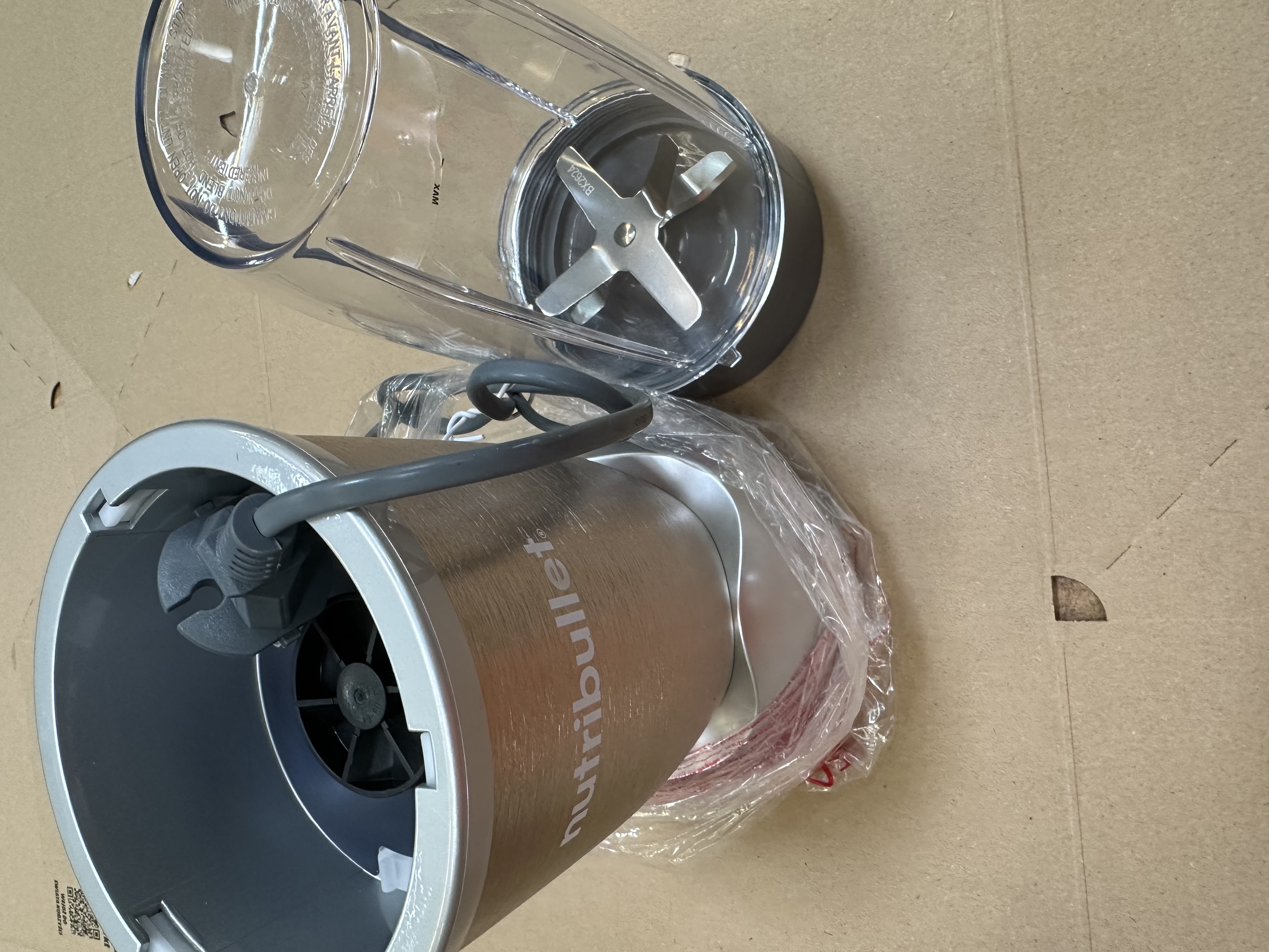 Blender Nutribullet, srebrna podstawa, przezroczysty plastikowy kubek i zespół ostrzy na kartonie.
