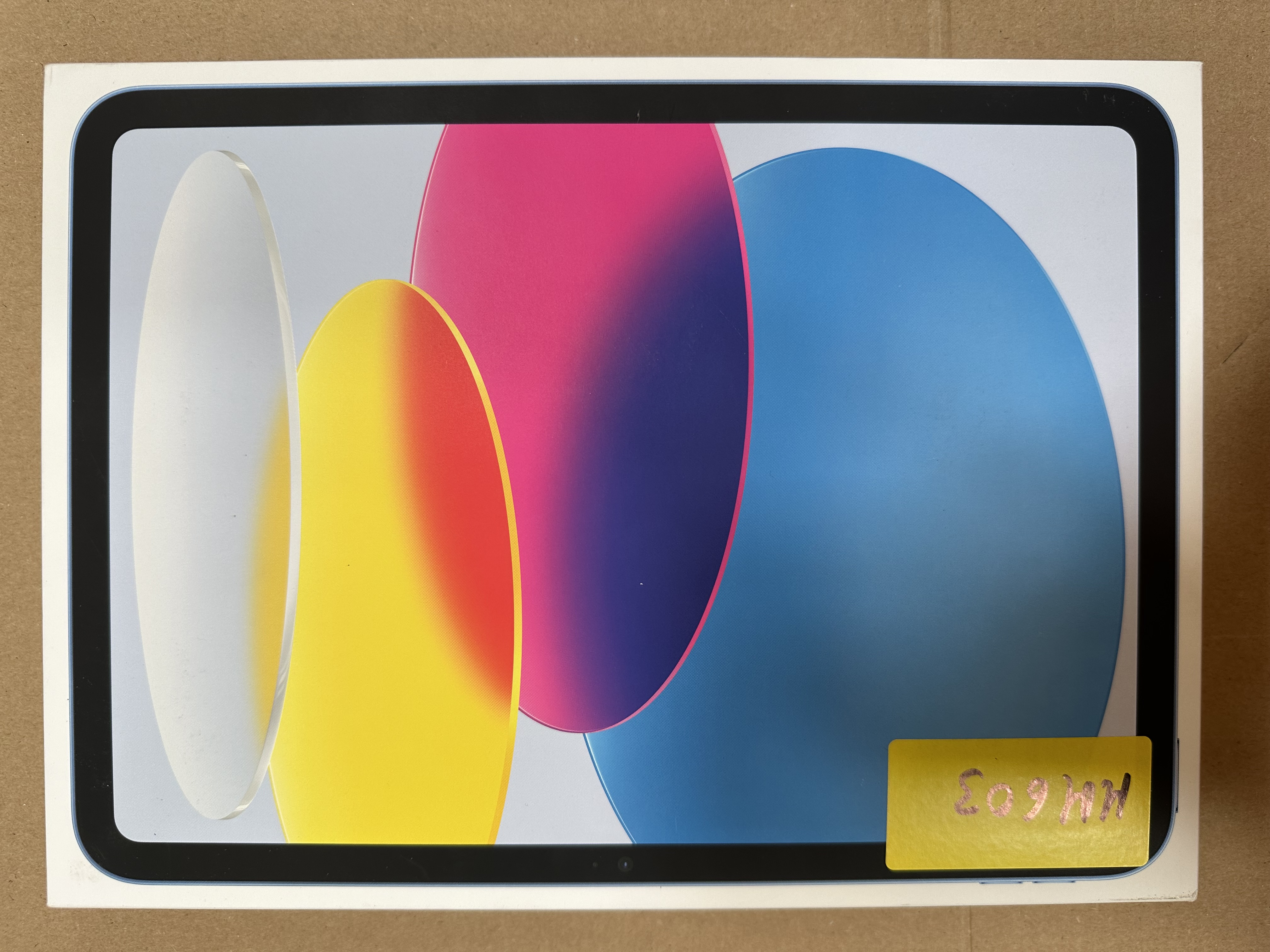 Pudełko z kolorowymi abstrakcyjnymi kształtami. Żółta naklejka z odręcznym tekstem znajduje się w prawym dolnym rogu.