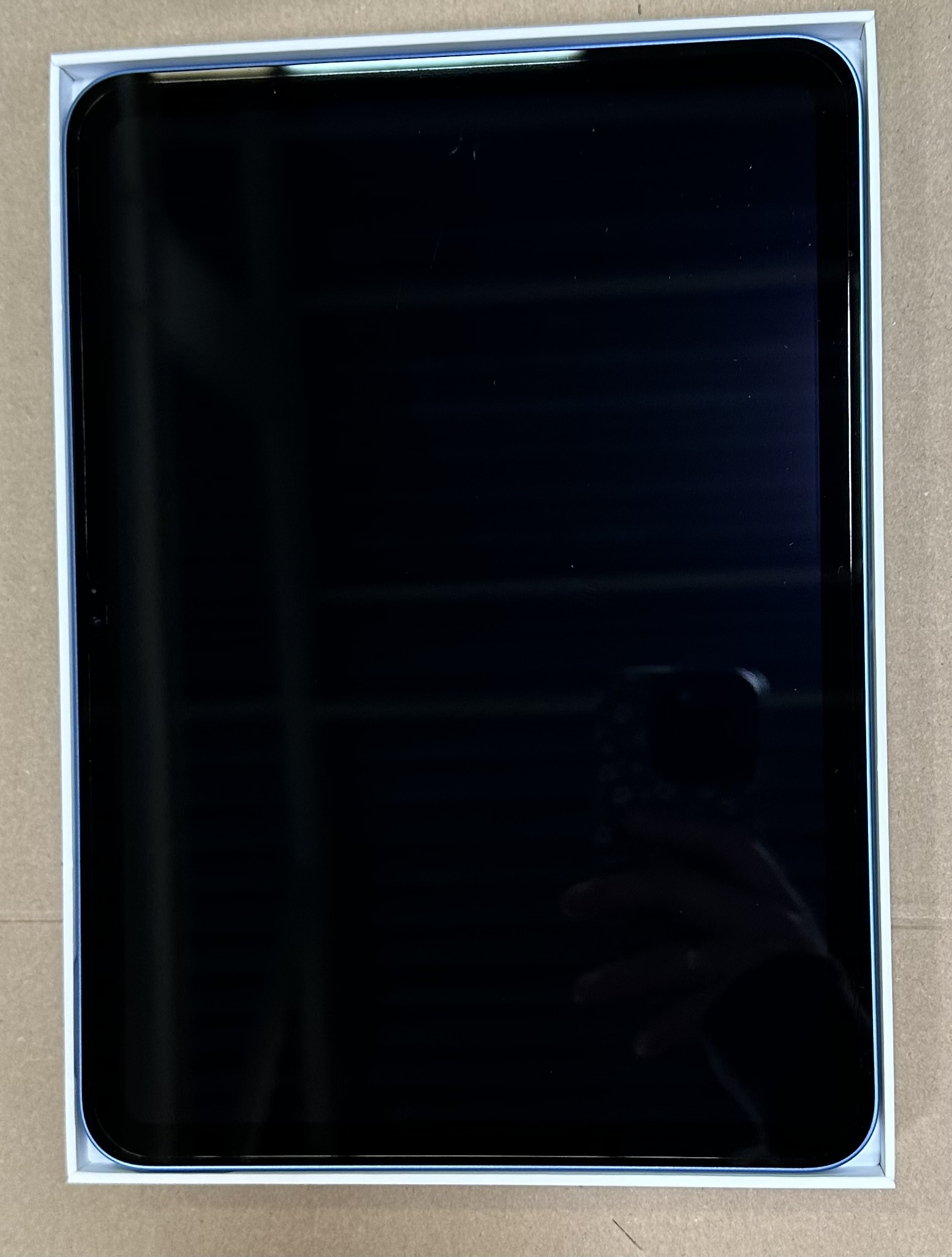 Czarny tablet w białym pudełku na brązowej powierzchni. Ekran odbija osobę z telefonem.