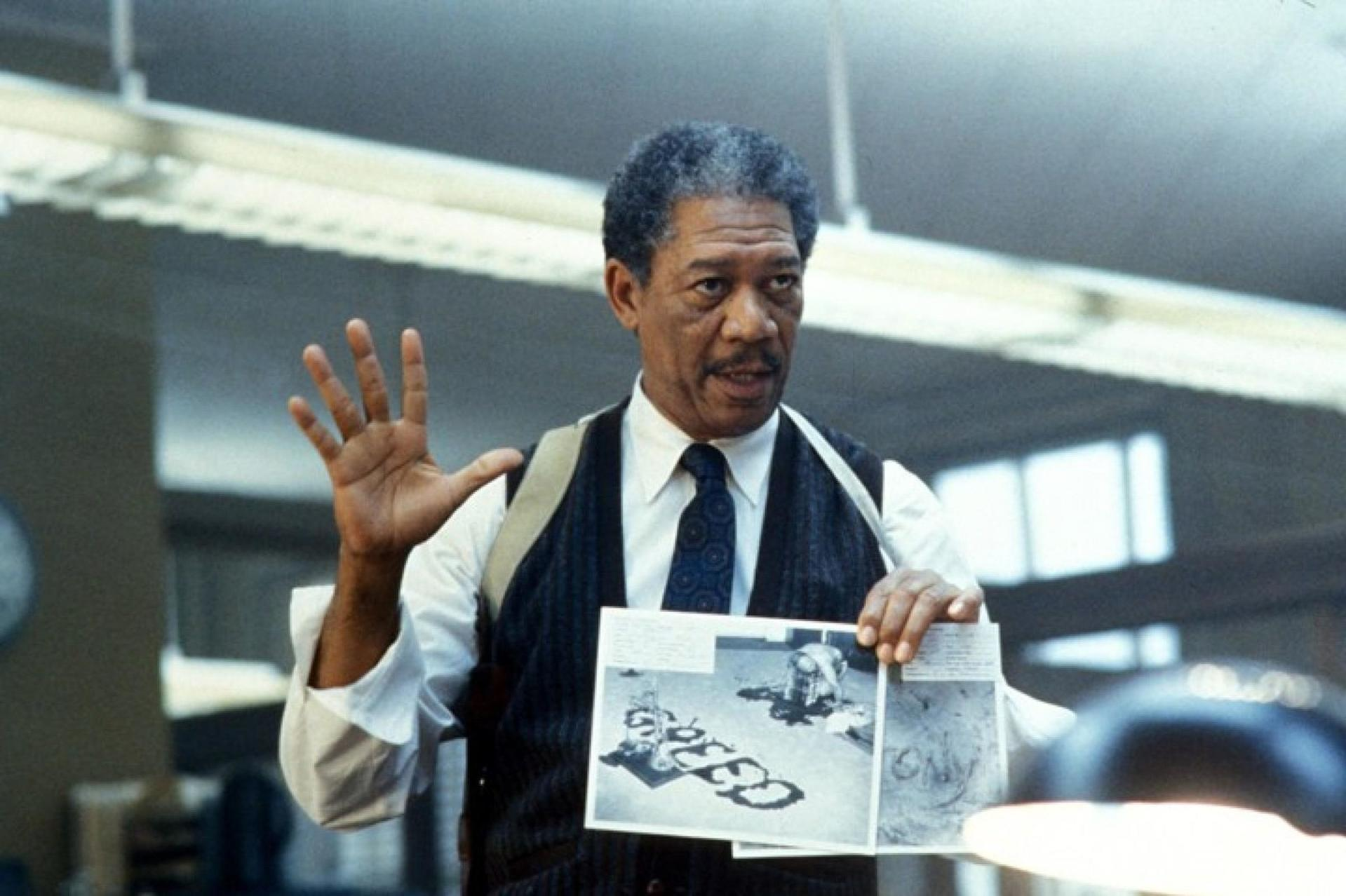Morgan Freeman hält Fotos, gestikuliert mit offener Hand, in einem Büro.