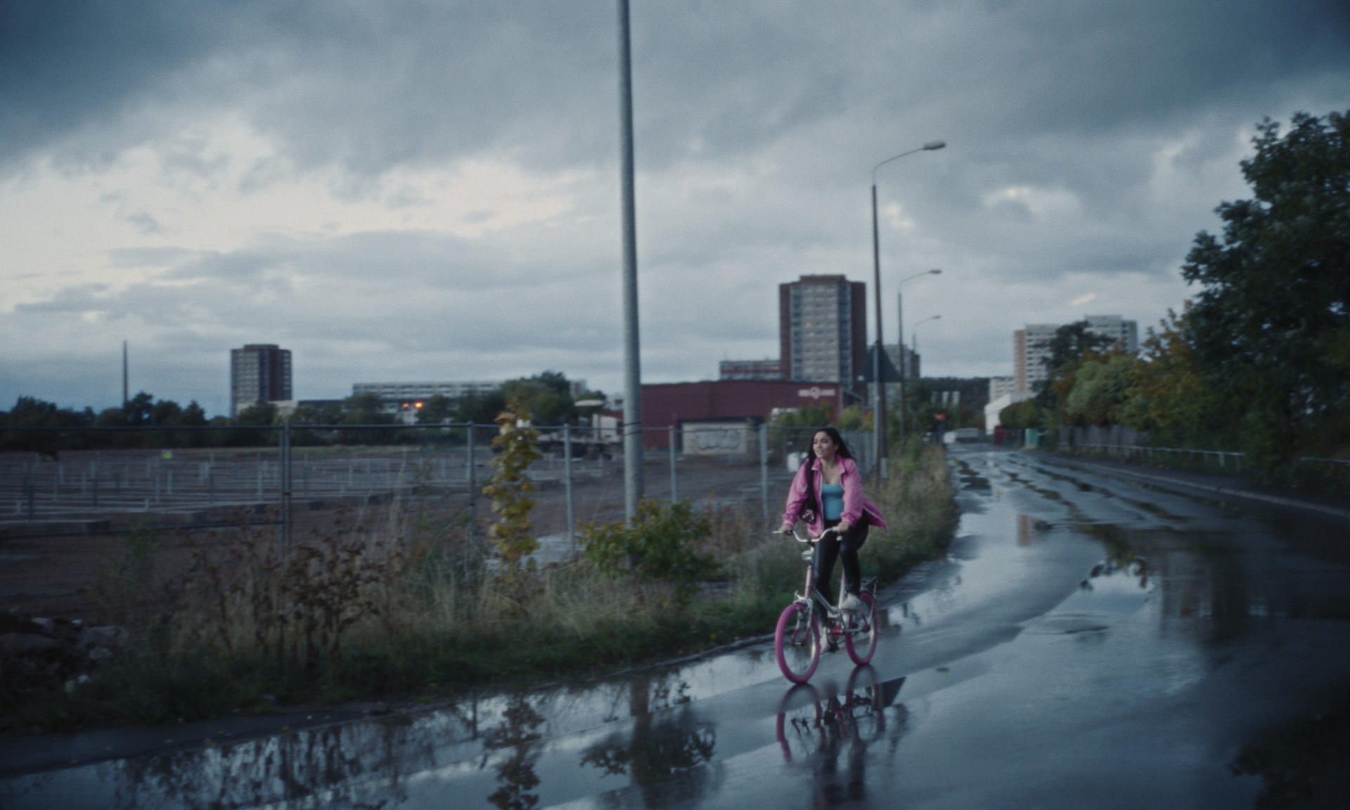 Eine Frau fährt mit einem pinken Fahrrad auf nasser Straße. Bewölkter Himmel, Industriegebäude im HG.