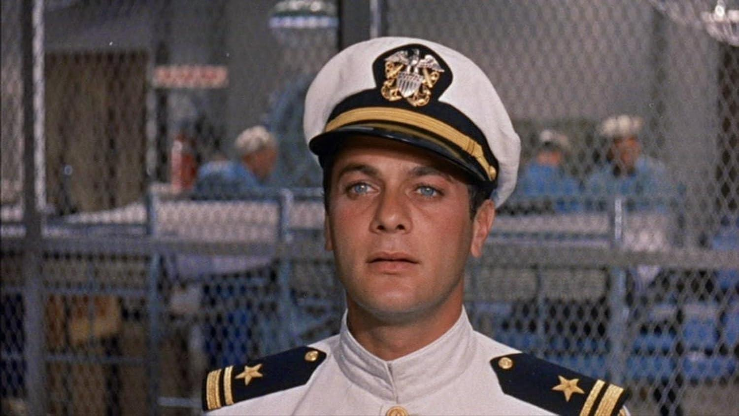 Mann in Marineuniform mit Hut, blickt nach oben, in einem Raum mit Etagenbetten, mit anderen Männern im Hintergrund.