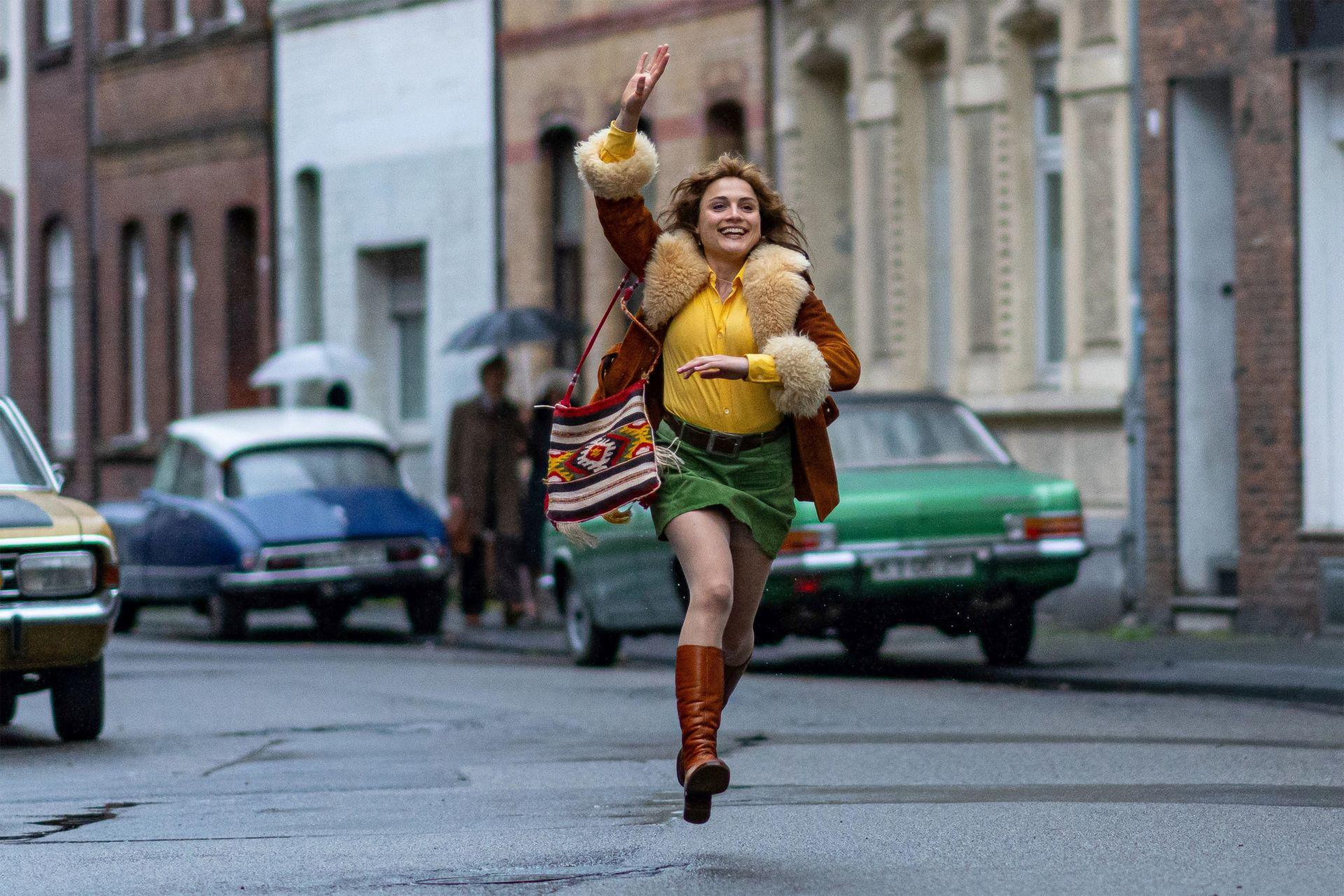 Eine Frau in Retro-Kleidung rennt eine Straße entlang und winkt mit der Hand. Autos stehen am Straßenrand.