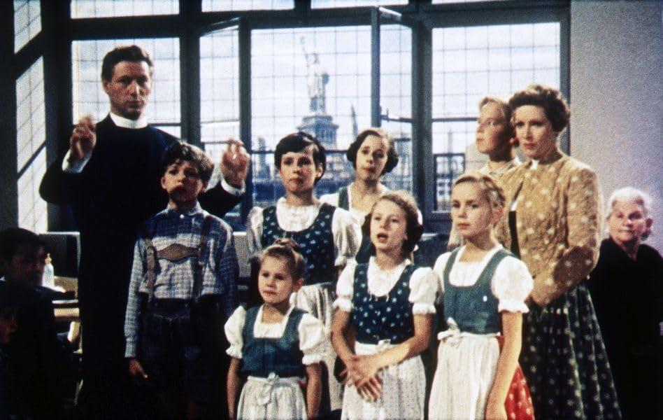 Eine Gruppe von Leuten, darunter Kinder, befinden sich in einem Raum, in dem ein Mann sie dirigiert.