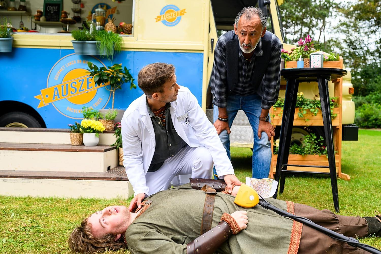 Ein Mann liegt auf Gras, andere um ihn herum. Ein Metalldetektor ist in seiner Nähe, und ein Foodtruck ist im Hintergrund.