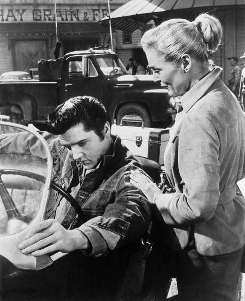 Elvis in einem Auto, blickt nach unten. Eine Frau berührt seine Schulter. Hintergrund: LKW, Gebäude.