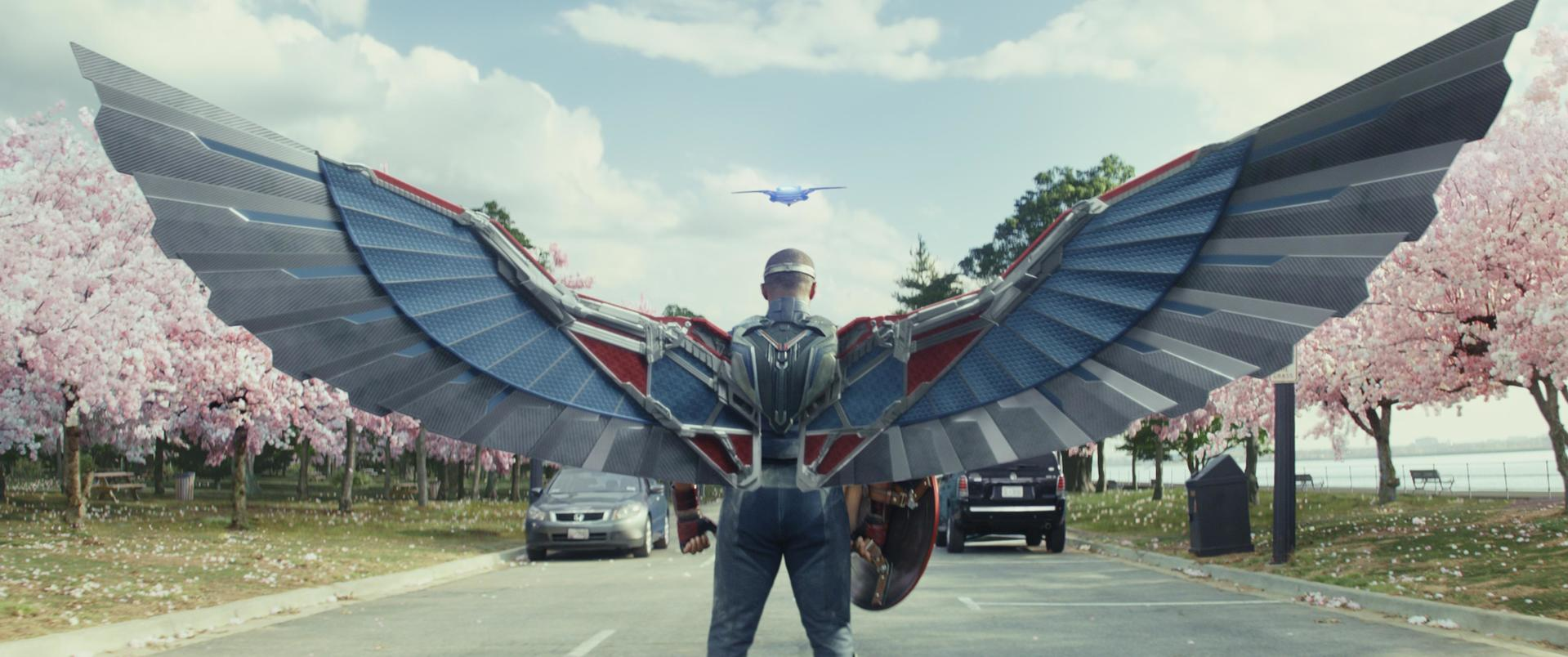 Ein Mann mit Flügeln steht in einem Park. Bäume und ein blaues Flugzeug im Hintergrund.