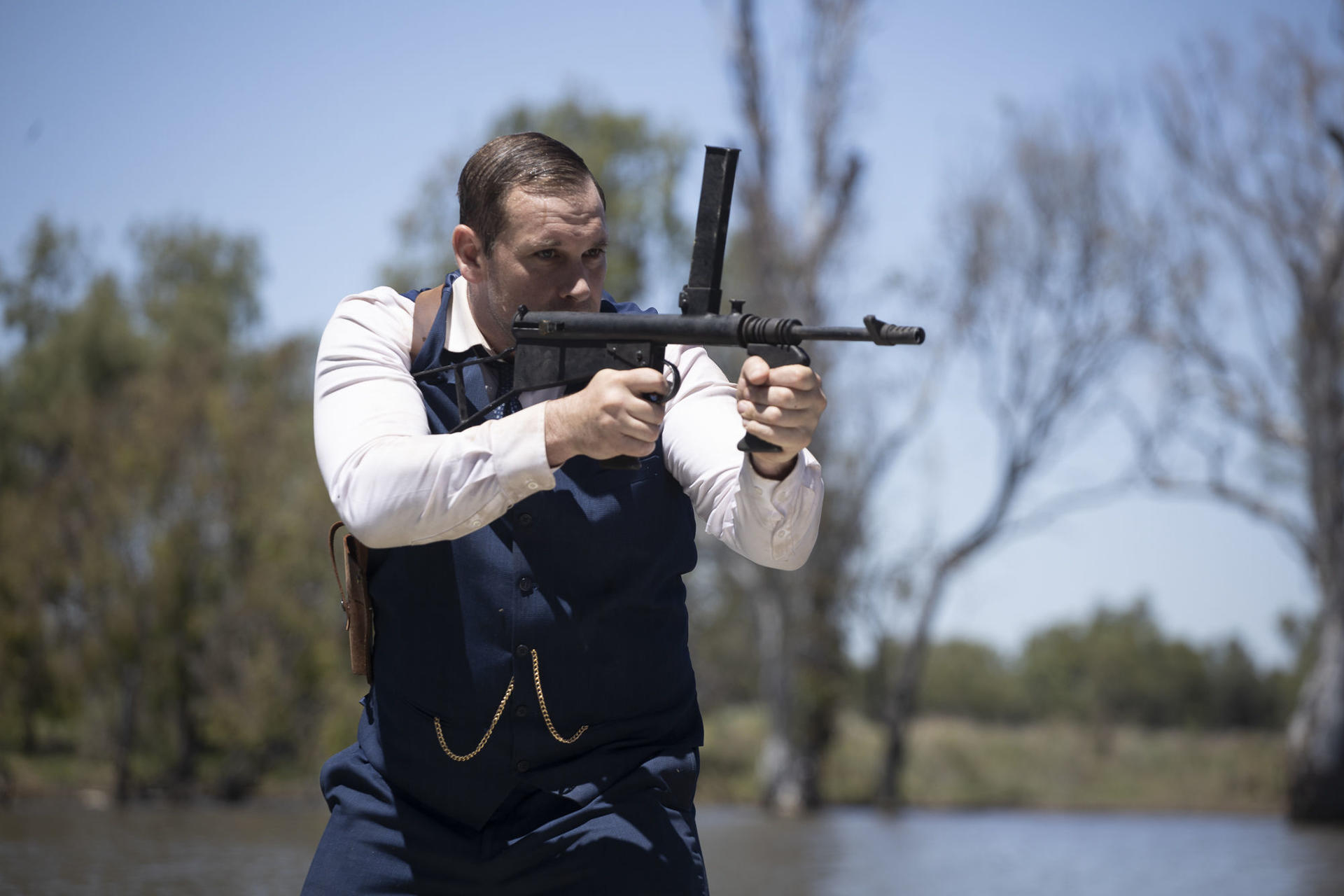Ein Mann zielt mit einem Maschinengewehr. Er trägt eine dunkle Weste und ein weißes Hemd. Ein Gewässer ist im Hintergrund.