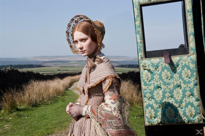 Eine Frau in historischer Kleidung steht auf einem Pfad und blickt weg. Grünes Feld und Hügel im Hintergrund.