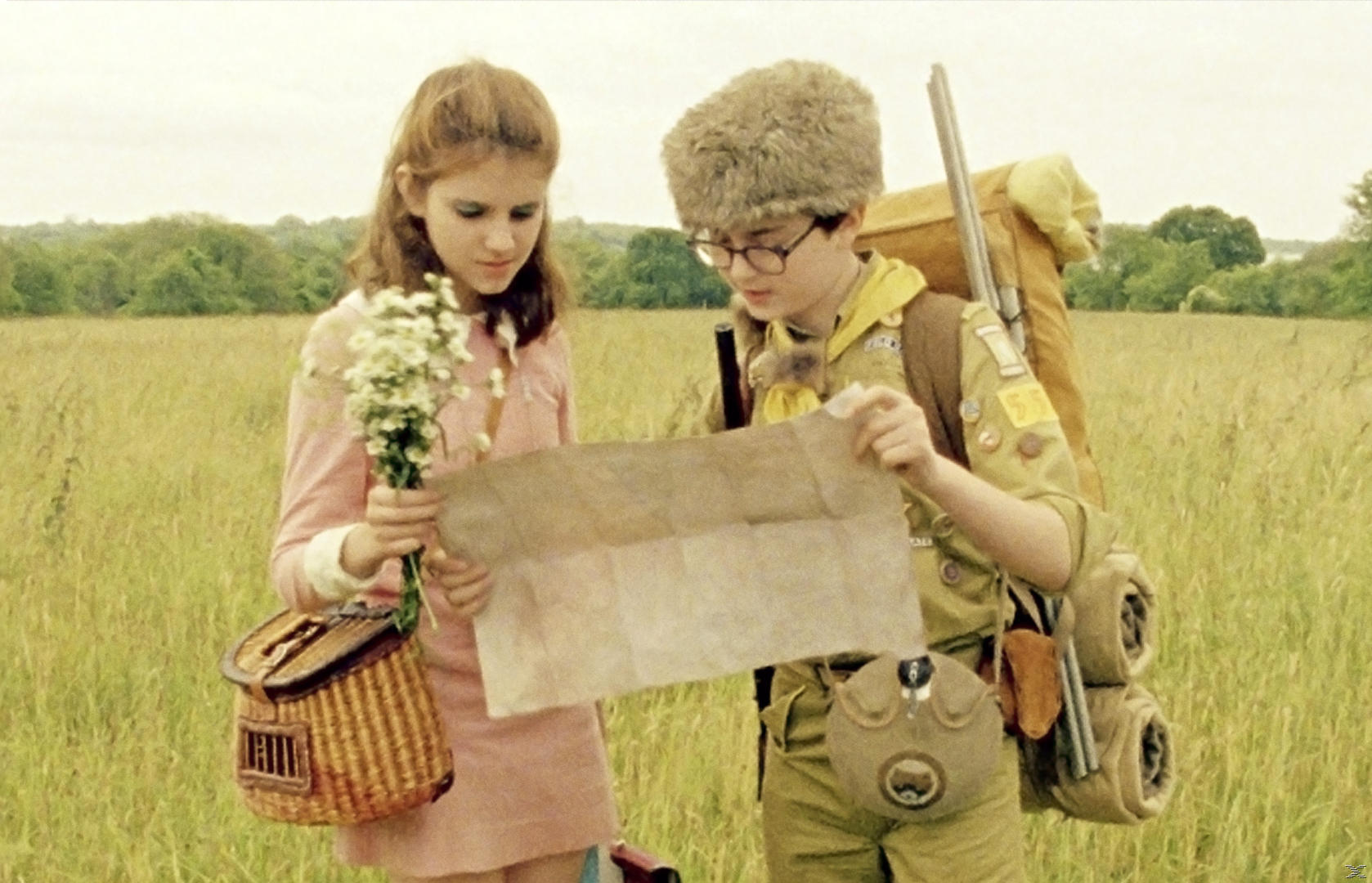 Zwei Kinder betrachten eine Karte auf einem Feld. Eines hält Blumen, das andere einen Rucksack.
