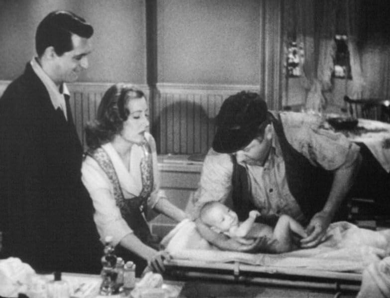 Schwarz-Weiß-Foto von zwei Männern und einer Frau, die ein Baby auf einem Tisch betrachten.