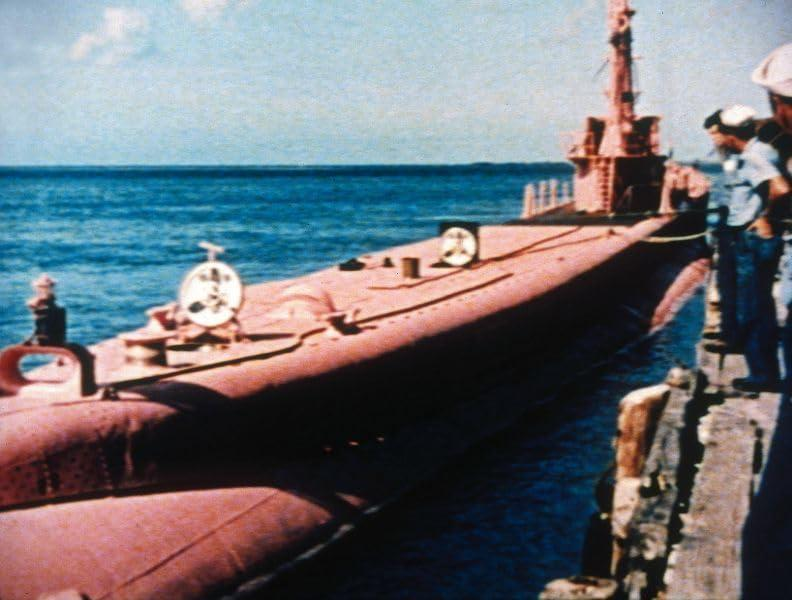 Ein rosa U-Boot liegt an einem Pier, Menschen stehen in der Nähe unter hellem Himmel.