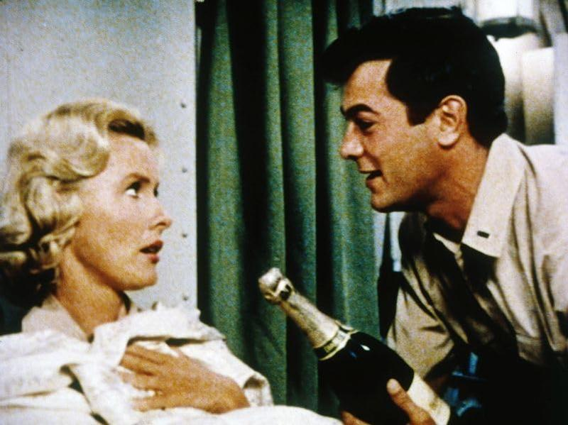 Mann bietet einer blonden Frau Champagner an. Sie ist in eine weiße Decke gehüllt.