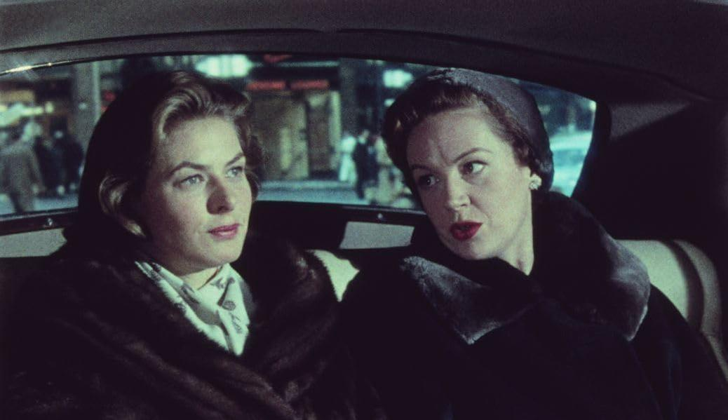 Zwei Frauen in einem Auto. Eine trägt einen Pelzmantel, die andere einen Hut. Sie schauen sich an.