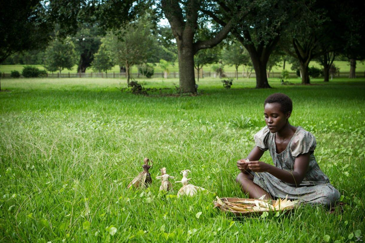 Eine Frau sitzt im Gras und macht Puppen. Drei Puppen sind neben ihr. Bäume und Himmel im Hintergrund.