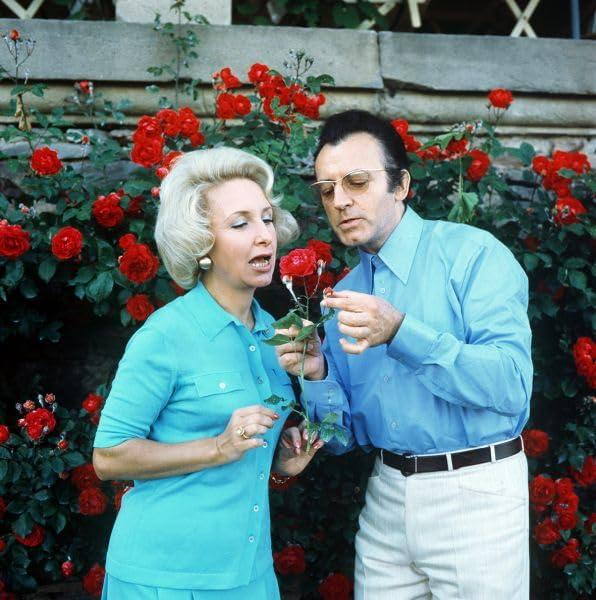 Ein Mann und eine Frau riechen im Freien an einem roten Rosenbusch. Der Mann trägt eine Brille.