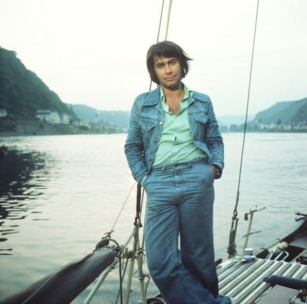 Ein Mann in einer Jeansjacke steht auf einem Boot und blickt in die Kamera. Hintergrund zeigt Fluss und Berge.