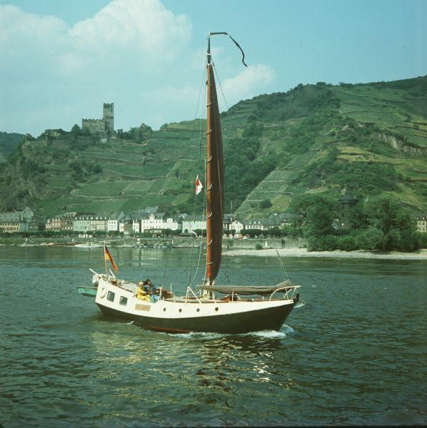 Ein Segelboot auf einem Fluss, mit grünen Hügeln im Hintergrund. Der Himmel ist blau mit weißen Wolken.