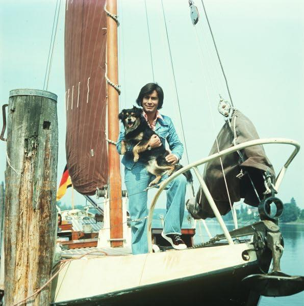 Ein Mann mit Hund auf einem Boot, lächelnd, mit der deutschen Flagge im Hintergrund.