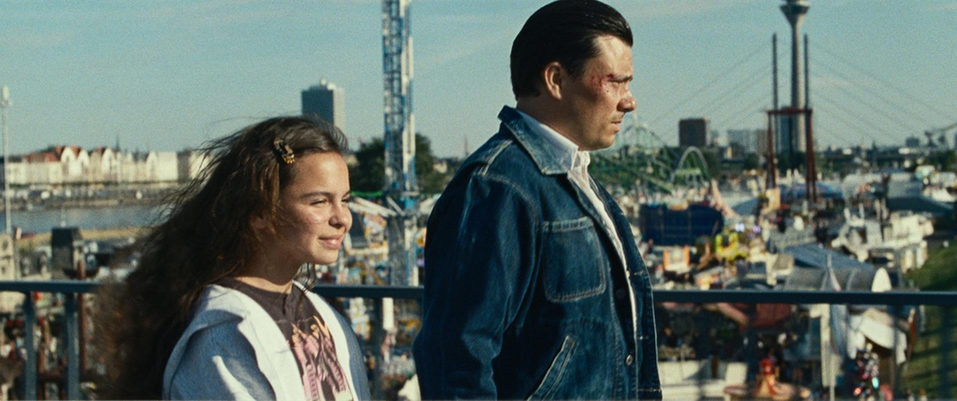 Zwei Personen auf einem Dach, ein Mädchen und ein Mann, mit Stadtblick im Hintergrund.