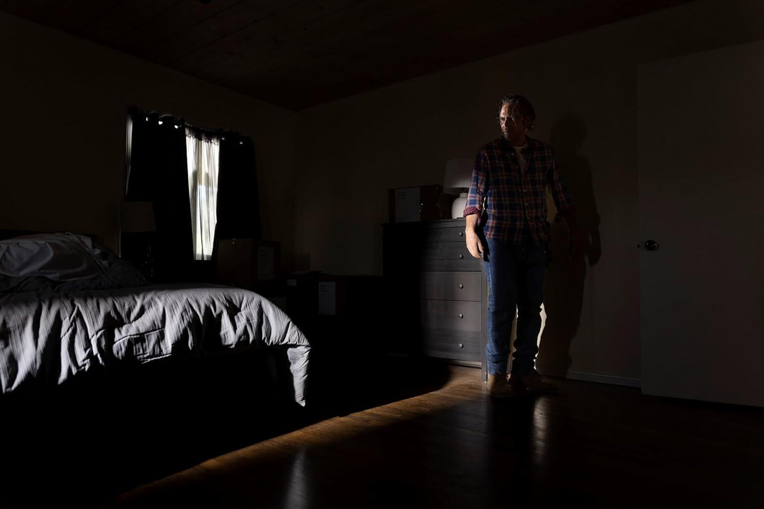 Ein Mann im karierten Hemd steht in einem schwach beleuchteten Raum. Ein Bett ist in der linken Ecke. Sonnenlicht scheint durch ein Fenster.
