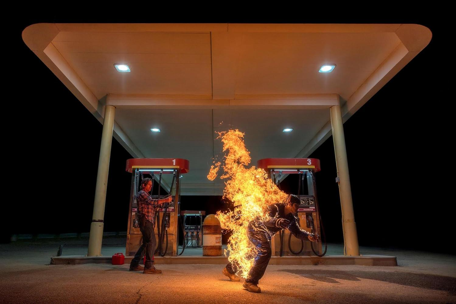 Ein Mann steht in Flammen an einer Tankstelle, ein anderer Mann zielt mit einer Waffe.