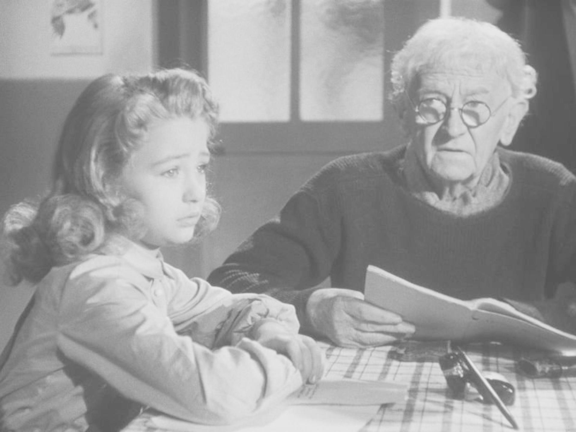 Ein junges Mädchen und ein älterer Mann sitzen an einem Tisch. Der Mann hält ein Papier, das Mädchen ist traurig.