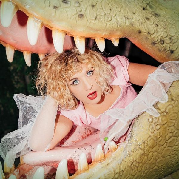 Eine Frau in einem rosa Kleid sitzt in einem großen, offenen Alligatormaul und sieht überrascht aus.