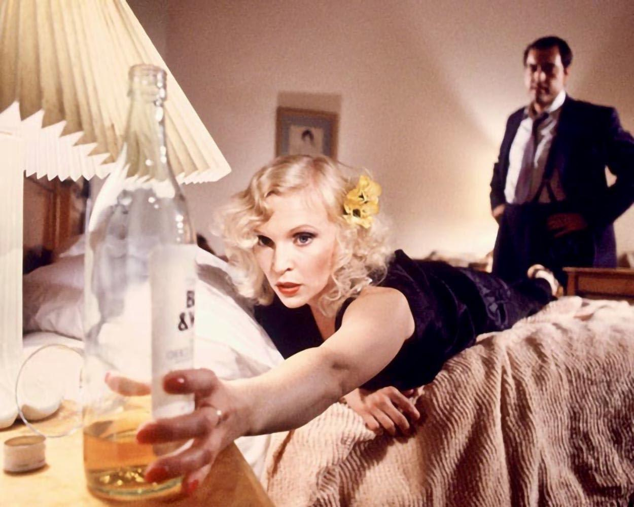 Eine Frau in einem schwarzen Kleid greift nach einer Flasche auf einem Tisch. Ein Mann steht hinter ihr.