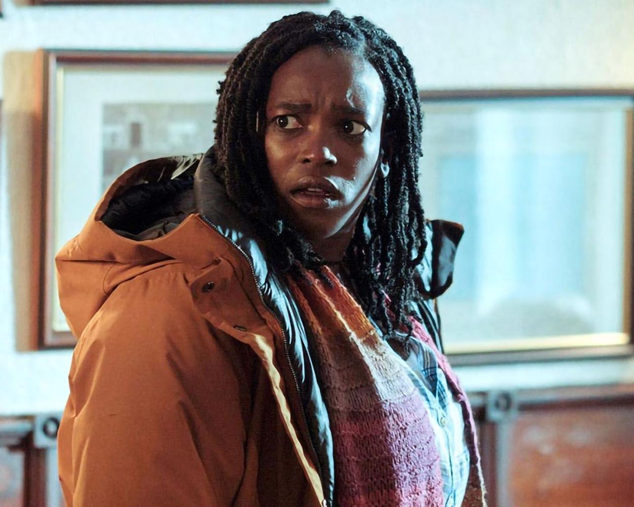 Eine Frau mit Dreadlocks, die besorgt aussieht. Sie trägt einen orangefarbenen Mantel und einen Schal.