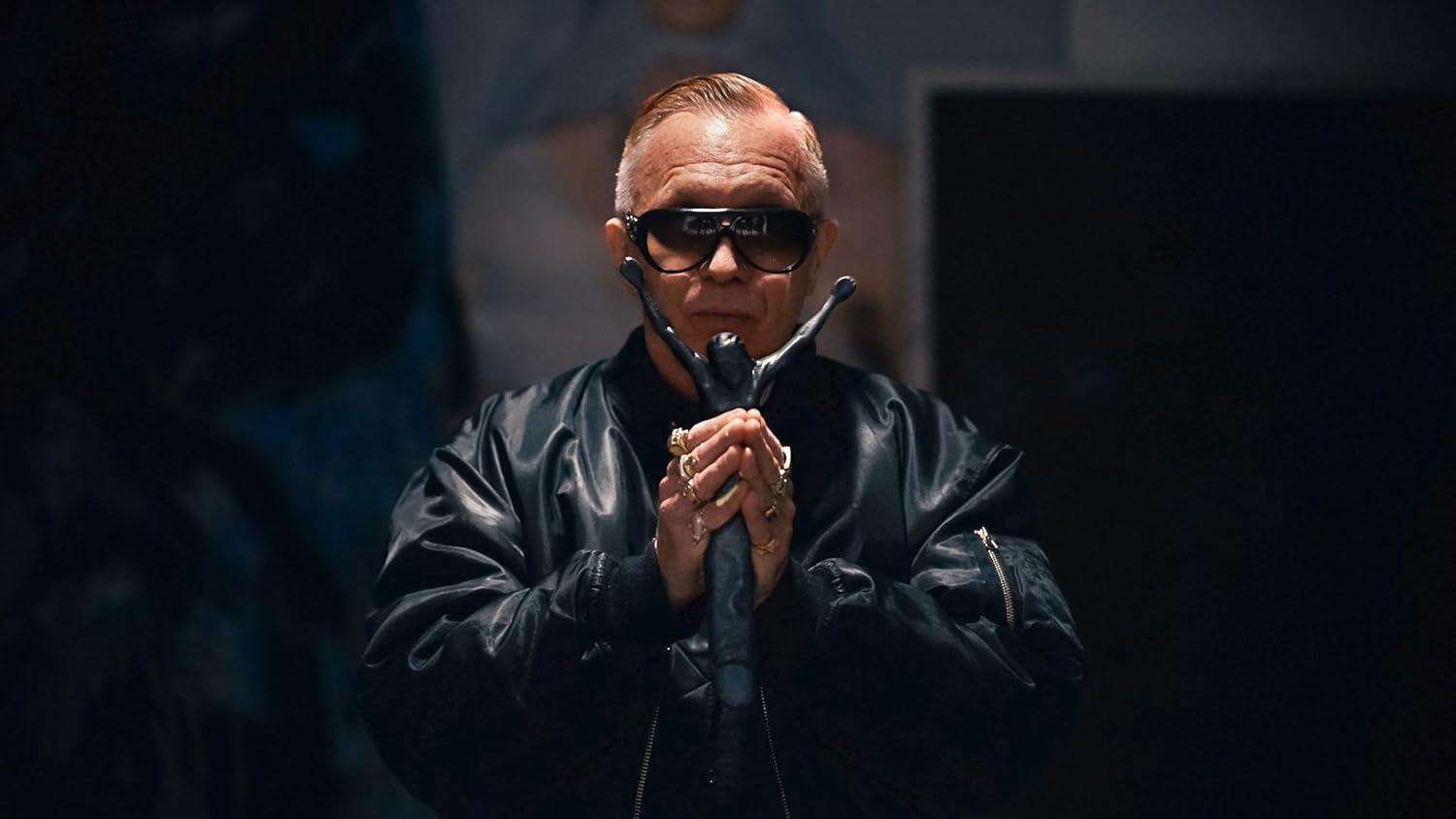Mann mit Sonnenbrille hält Stab, trägt schwarze Jacke, Ringe und einen ernsten Gesichtsausdruck.