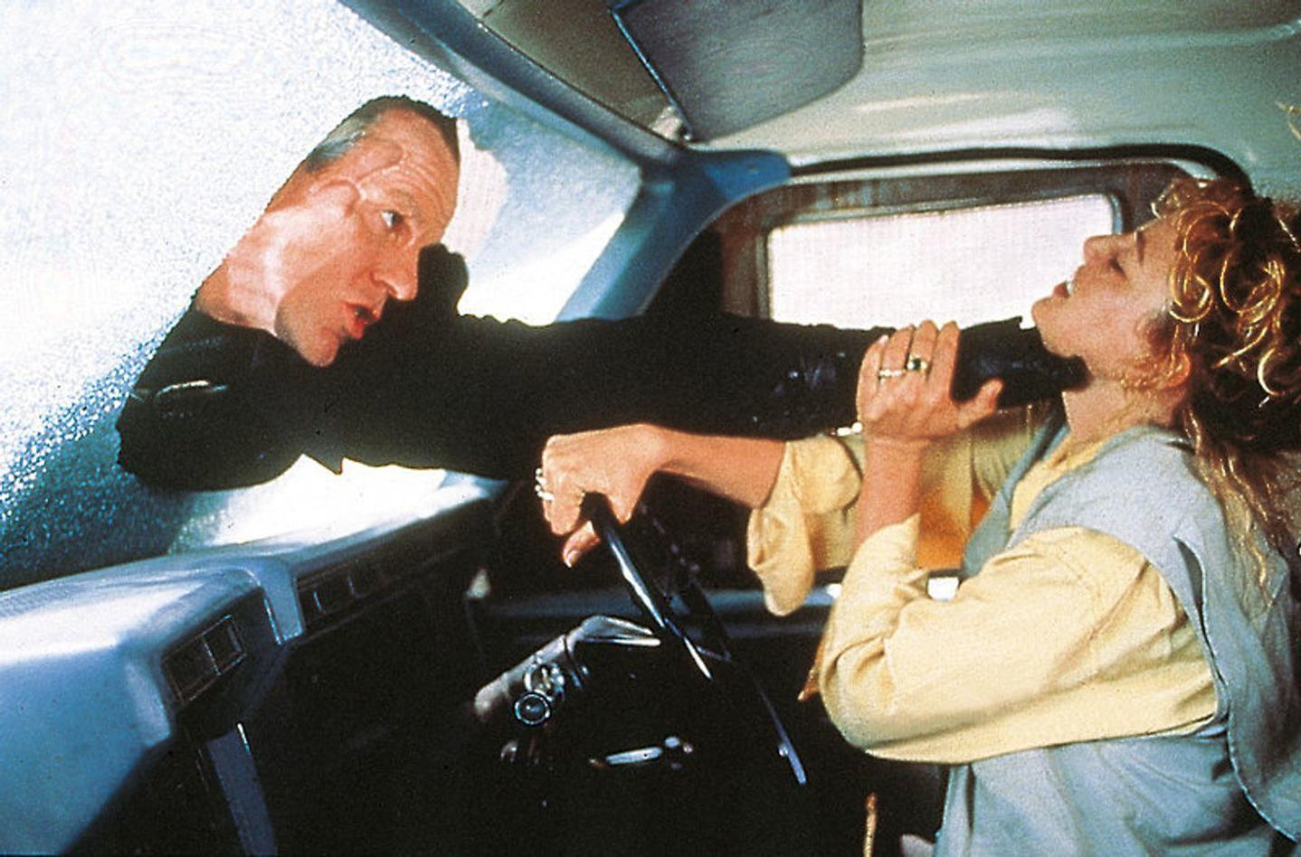 Ein Mann greift durch ein Autofenster und richtet eine Waffe auf eine Frau im Auto.