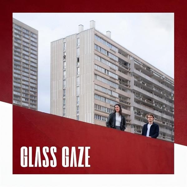 Zwei Personen stehen auf einer roten Mauer mit Apartmentgebäuden im Hintergrund. Text: GLASS GAZE.