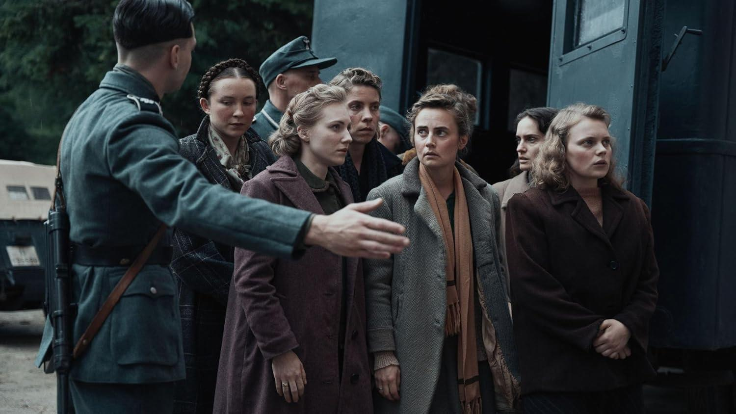 Szene aus dem Zweiten Weltkrieg: Soldaten, Frauen in Mänteln, ein Zugwagen. Ein Soldat deutet auf die Frauen.