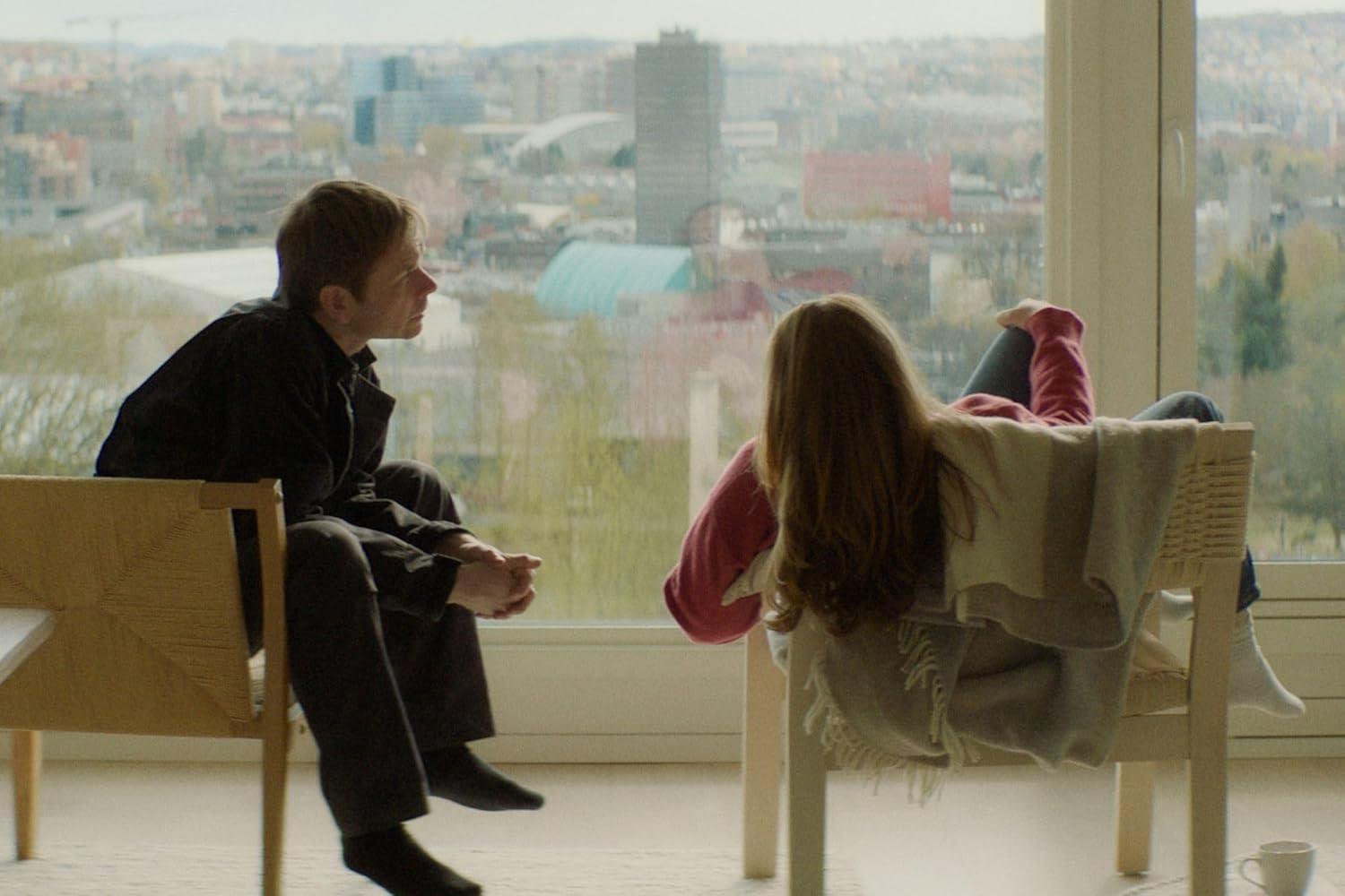 Ein Mann und eine Frau in einem Raum mit Stadtblick. Sie sitzen auf Stühlen und unterhalten sich.