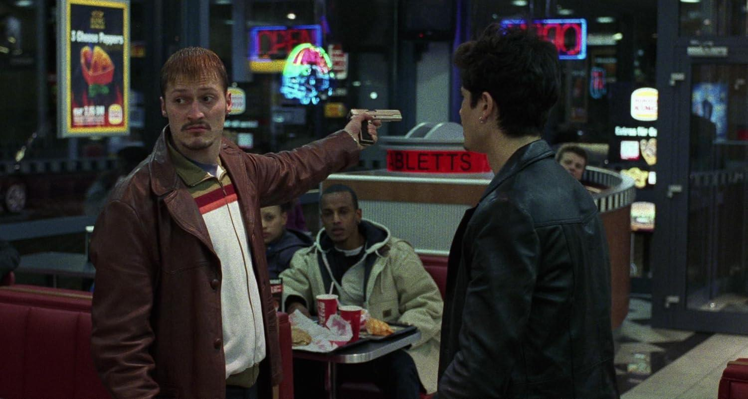 Mann in Lederjacke richtet Waffe auf anderen Mann in Diner, andere schauen zu.