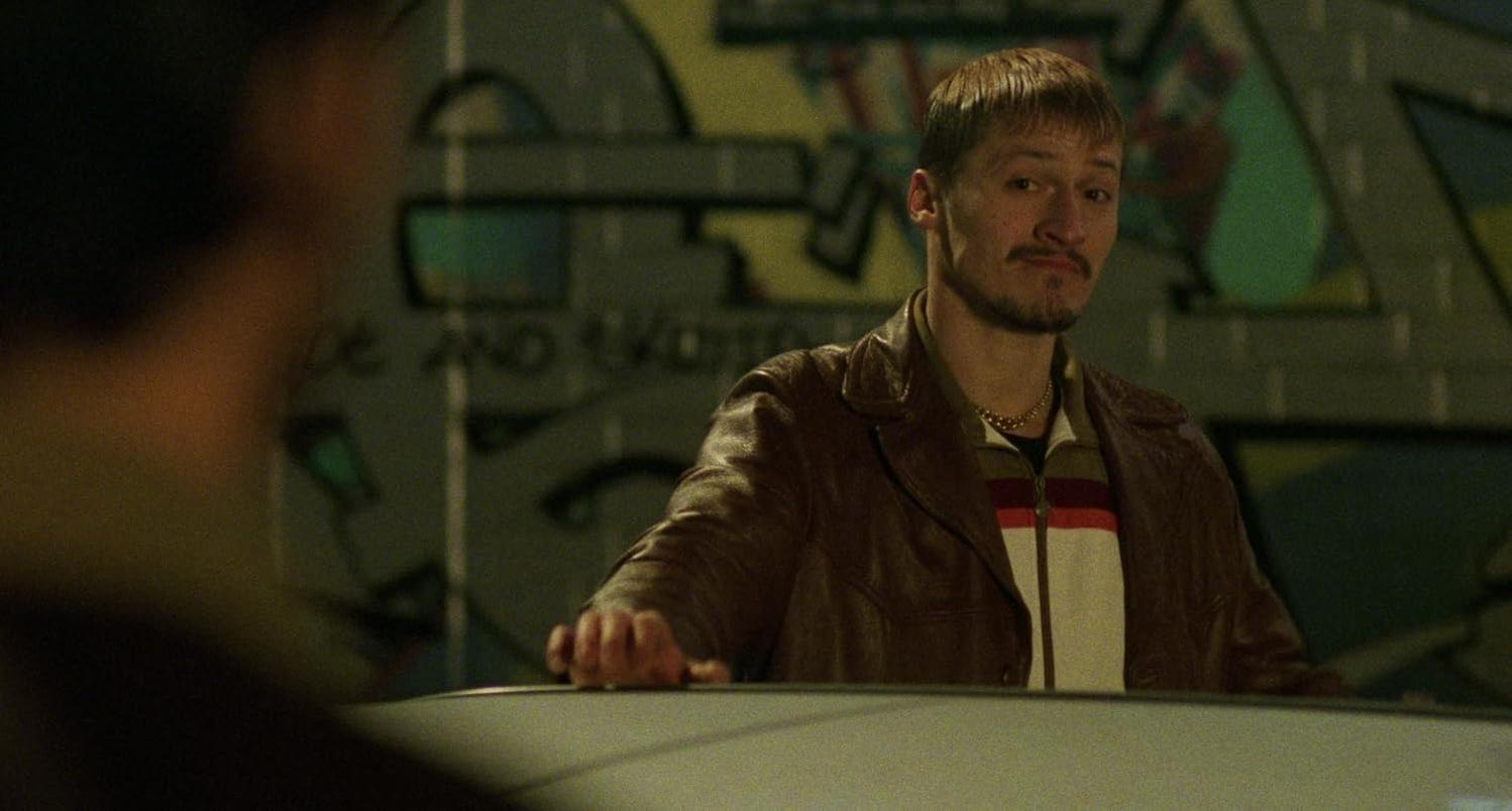 Ein Mann mit einer braunen Lederjacke und ein Mann im Hintergrund vor einer Graffiti-Wand.