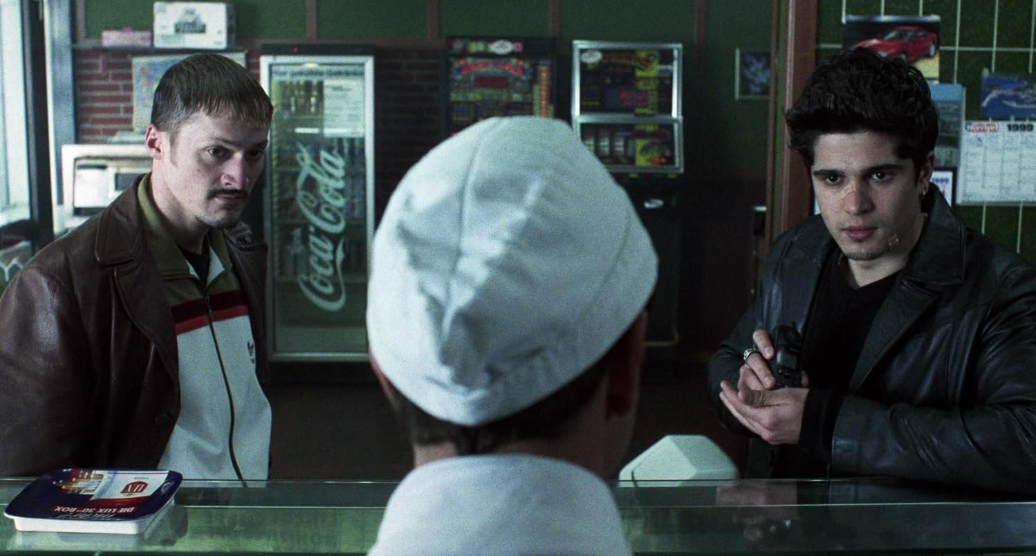 Szene aus einem Film. Drei Männer sind in einem Laden. Einer mit Hut steht hinter der Theke.